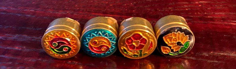 May include: Five small, round, gold-colored metal containers with colorful enamel designs. The designs include a yin-yang symbol with a sun, a yin-yang symbol with a sun, a red flower, and a yellow flower.