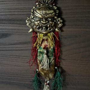 May include: A gold-colored metal pendant with a fierce-looking face, surrounded by red, yellow, and green fabric tassels. The pendant is hanging from a string.