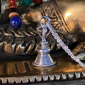 May include: Silver bell pendant with a detailed design and a silver chain. The bell is hanging on a gold surface.
