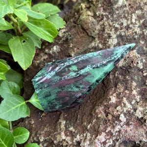 May include: A polished, pointed crystal tower featuring a mottled pattern of deep forest green and vibrant magenta-purple. The stone has a smooth, reflective surface with visible natural mineral inclusions and sharp, angular edges.