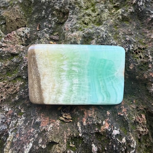 May include: A rectangular, polished stone with a gradient of colors. The stone transitions from a light brown on one end, through cream and light green, to a vibrant turquoise on the other. The stone's surface has a subtle, natural texture.