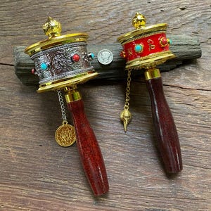May include: Two gold prayer wheels with red and turquoise accents. The wheels are attached to wooden handles and have a chain with a seashell charm.