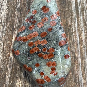Pu&ograve; includere: Una pietra levigata di forma triangolare caratterizzata da una base liscia color verde salvia con numerose macchie distinte, arrotondate e bruno-rossastre sparse sulla superficie, che creano un motivo naturale punteggiato.