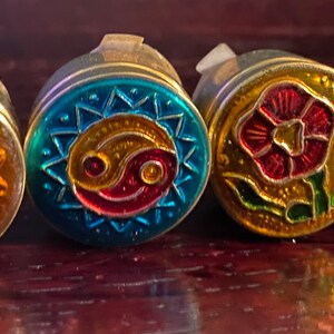 May include: Five round, decorative, metal trinket boxes with different designs. The boxes are gold, blue, gold, black, and gold with various colors and patterns. The designs include a yin yang symbol, a flower, and a lotus flower.