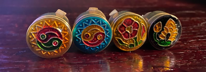 May include: Five round, decorative, metal trinket boxes with different designs. The boxes are gold, blue, gold, black, and gold with various colors and patterns. The designs include a yin yang symbol, a flower, and a lotus flower.