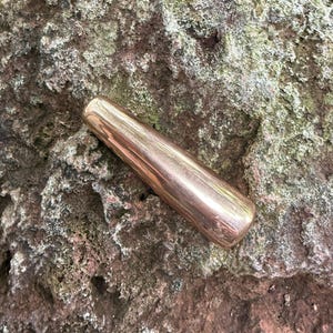 May include: A smooth, polished, cylindrical object with a tapered end, featuring a metallic copper-toned finish. The item has a solid, uniform appearance with a reflective surface that catches light along its length, showing a warm, reddish-brown hue.