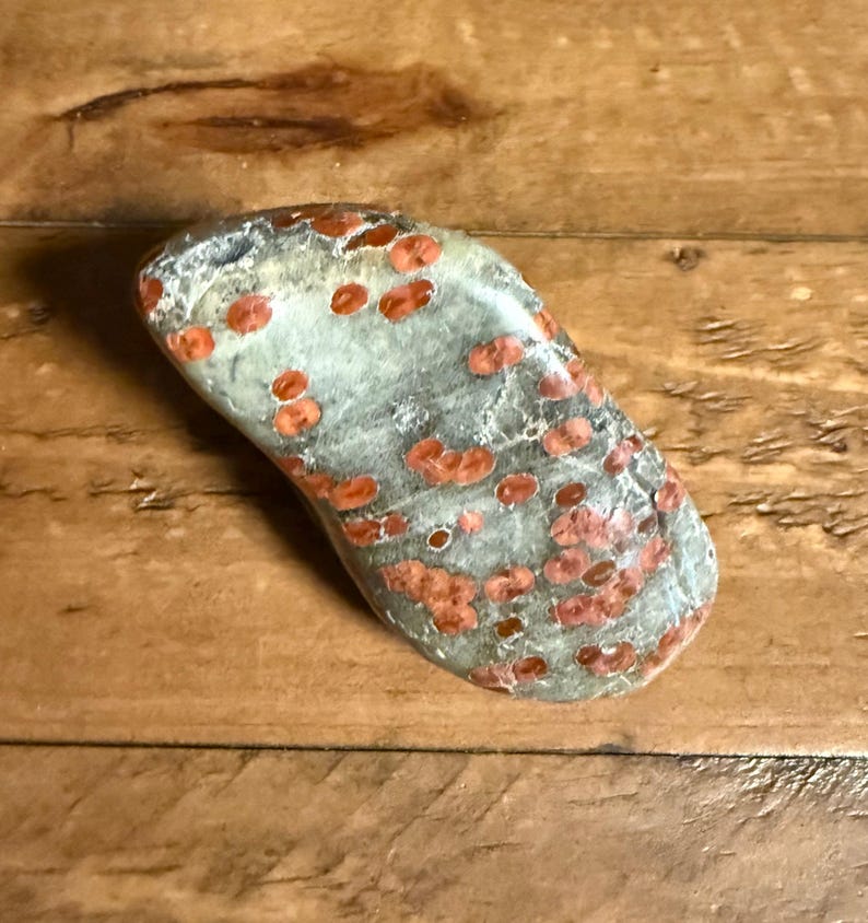 Pu&ograve; includere: Una pietra levigata e liscia a forma di rene, caratterizzata da una base di colore grigio chiaro e verde salvia con numerose macchie rotonde distinte di colore rosso-arancio sparse sulla superficie in un motivo naturale irregolare.