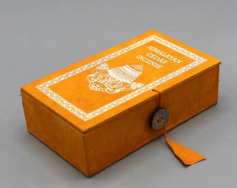 Può includere: Una scatola gialla con un design bianco e il testo "Himalayan Cedar Incense". La scatola ha una chiusura a cordoncino con un bottone di legno.