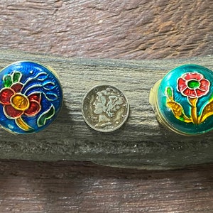 May include: Five small round trinket boxes with floral designs. The boxes are painted in shades of blue, red, yellow, and green. The boxes are arranged in a row on a piece of driftwood.