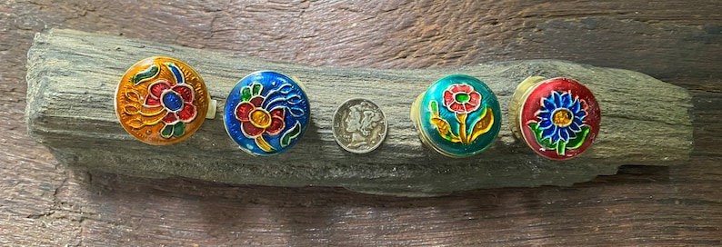 May include: Five small round trinket boxes with floral designs. The boxes are painted in shades of blue, red, yellow, and green. The boxes are arranged in a row on a piece of driftwood.
