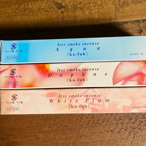 May include: Three rectangular boxes of Japanese incense are stacked. The top box is light blue with the word "Aqua" and "less smoke incense" printed on it. The middle box is pink with the word "Daphne" and "less smoke incense" printed on it. The bottom box is pink with the words "White Plum" and "less smoke incense" printed on it.