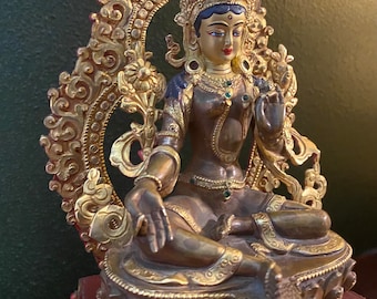 May include: A gold-colored statue of a deity with a crown and ornate halo. The statue is sitting on a lotus throne with intricate details. The deity has a serene expression and is adorned with jewels.