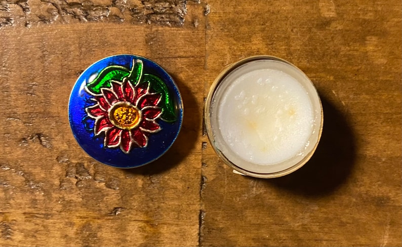 May include: A small, round, blue tin with a red, yellow, and green flower design on the lid. The tin is open and the inside is empty.