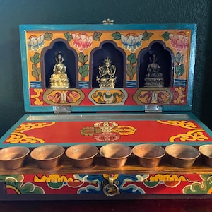 May include: A wooden box with a red, blue, and yellow painted design. The box is open and has three small golden statues inside. The box also has eight small brass bowls on the bottom.