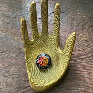May include: A gold metal hand-shaped ring holder with an intricate design. A black and gold ring with a red and yellow yin-yang symbol is displayed on the holder.