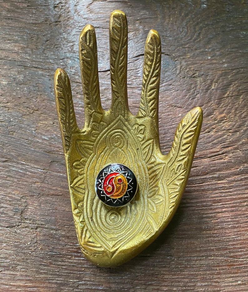 May include: A gold metal hand-shaped ring holder with an intricate design. A black and gold ring with a red and yellow yin-yang symbol is displayed on the holder.