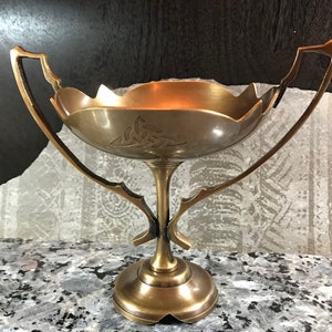 May include: A gold-toned metal bowl with two handles and a Celtic knot design. The bowl sits on a pedestal base.
