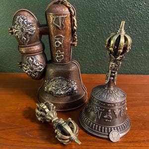 May include: A set of three Tibetan Buddhist ritual objects: a hand-held bell, a prayer wheel, and a vajra. The bell is made of metal and has a silver finish. The prayer wheel is made of copper and has a silver finish. The vajra is made of metal and has a silver finish.