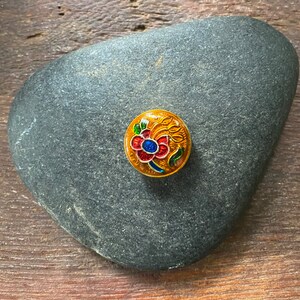 May include: A round, gold-colored button with a colorful floral design. The flower is red, blue, and green with yellow accents. The button is on a gray rock.