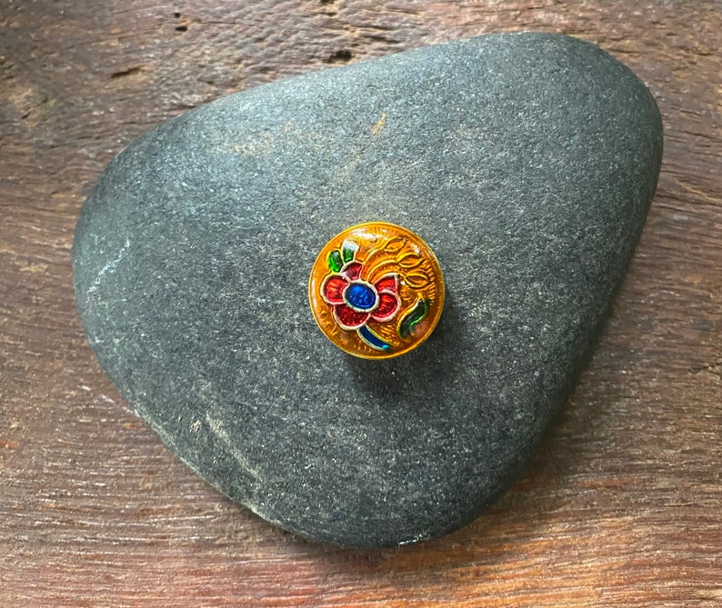 May include: A round, gold-colored button with a colorful floral design. The flower is red, blue, and green with yellow accents. The button is on a gray rock.