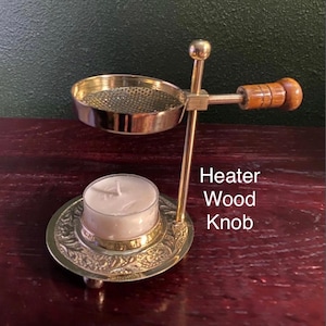 May include: A brass tea light candle heater featuring a circular base with an embossed floral pattern, a vertical support rod, and a top bowl with a mesh screen. A wooden knob is attached to the side of the bowl. The text Heater Wood Knob is visible.