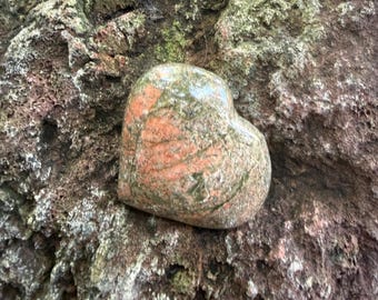 Può includere: Una pietra a forma di cuore con una superficie lucida caratterizzata da un motivo maculato di inclusioni minerali color rosa salmone, verde oliva e grigio. La pietra ha una consistenza liscia e arrotondata con variazioni naturali di colore.