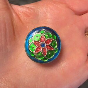 May include: A round, blue, green, and red enamel button with a flower design. The flower has five petals and a blue center.