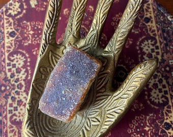 May include: A gold metal hand-shaped dish with intricate details holds a small, rectangular block of brown and white resin. The dish is on a red and gold patterned surface.