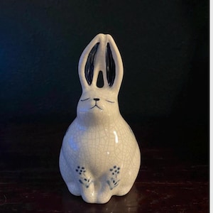 May include: A ceramic bunny figurine with a white crackle glaze. The bunny has long ears with dark blue interiors, closed eyes, and small blue floral accents. The figurine is sitting upright.