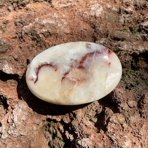 May include: A white and red agate palm stone with a smooth, polished surface. The stone has a swirling pattern of red and white.