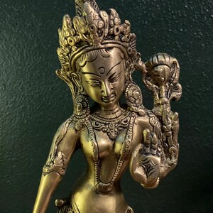May include: A gold-colored metal statue of a deity with a crown and intricate details. The statue is holding a lotus flower in its right hand.