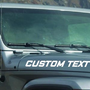 May include: A gray Jeep Wrangler with a white windshield banner that says "CUSTOM TEXT".