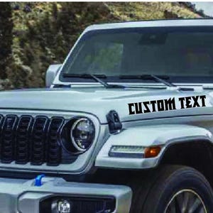 May include: A silver Jeep Wrangler with a black grille and black custom text decal on the hood that says "CUSTOM TEXT".