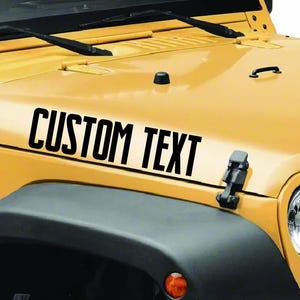 May include: A yellow vehicle hood with the words "CUSTOM TEXT" in bold, black letters. The hood has black windshield wipers, a black handle, and a black latch. The vehicle's front bumper and headlight are visible.