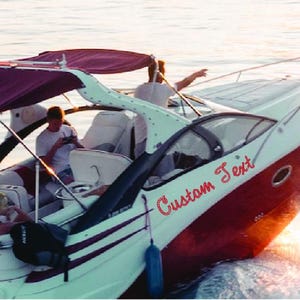 May include: A white and red motorboat with a burgundy canopy is cruising on the water. The boat has the words "Custom Text" in red script on the side. Several people are on board.