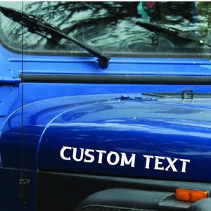 May include: A blue Jeep Wrangler with a white vinyl decal that says "CUSTOM TEXT" on the hood.