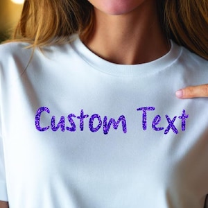 May include: White t-shirt with the words "Custom Text" in sparkly purple lettering. The t-shirt is being pointed at by a person's finger. The shirt is made of a soft material.