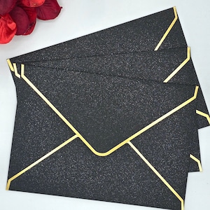 May include: A stack of black envelopes with gold glitter and gold trim. The envelopes are folded and ready to be used for sending letters or cards.