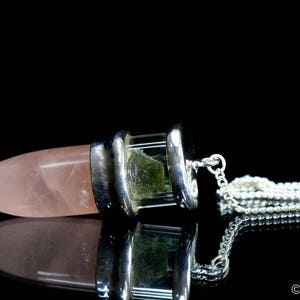 May include: A silver and rose quartz pendulum with a green crystal encased in a clear glass cylinder. The pendulum is attached to a silver chain with a clear quartz crystal at the end.