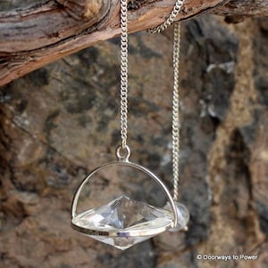 Puede incluir: Una cadena de plata con un péndulo de cristal transparente colgando de ella. El péndulo tiene forma de diamante y está suspendido de un anillo de plata. La cadena está colgando de una rama.