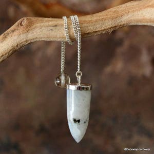 Può includere: Un pendolo in pietra di luna bianca con una catena d'argento e un cristallo di quarzo trasparente in cima. Il pendolo è appeso a un ramo di legno.