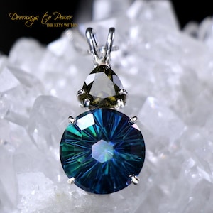 May include: A silver pendant with a round blue gemstone and a smaller, teardrop-shaped green gemstone. The pendant is set on a white background.