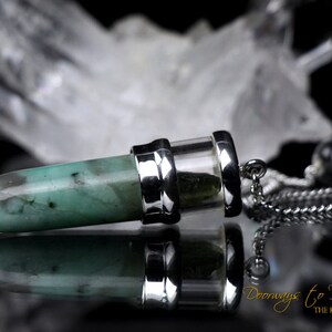 May include: A green emerald crystal point pendant with a silver casing and a silver chain. The pendant is hanging from a silver chain. The background is a blurry image of a white crystal.