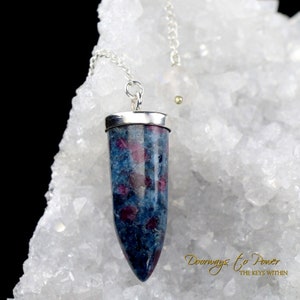 May include: A blue and red gemstone pendulum with a silver chain and accents. The pendulum is cylindrical with a pointed end and is suspended against a white, crystalline background. The text "Doorways to Power" is visible.