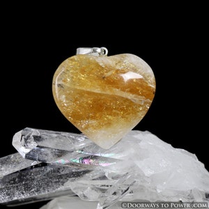 May include: A heart-shaped golden citrine pendant with a silver bail. The pendant is sitting on a bed of clear quartz crystals.