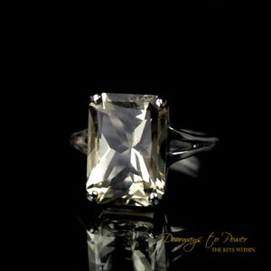 May include: A silver ring with a large, clear, rectangular-shaped gemstone. The gemstone has a slight yellow hue.