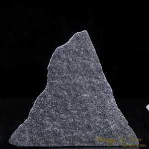 May include: A dark gray, triangular-shaped stone with a rough, textured surface. The stone has a jagged edge and a natural, organic appearance. The background is black, highlighting the stone's shape and texture.