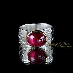 May include: A wide silver band ring featuring an oval-shaped, deep red gemstone set in a gold-toned bezel. The silver band has an embossed, textured pattern across its surface. The text "Doorways to Power THE KEYS WITHIN" appears in gold.