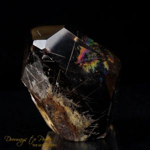 May include: A black, clear, and iridescent crystal with a point at the top. The crystal has many thin, white lines running through it. The crystal is on a black background.