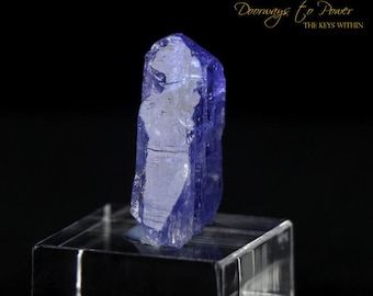 May include: A deep blue, translucent crystal with a pointed tip, sitting on a clear glass display stand. The crystal has a natural, rough texture and is a beautiful example of a gemstone.